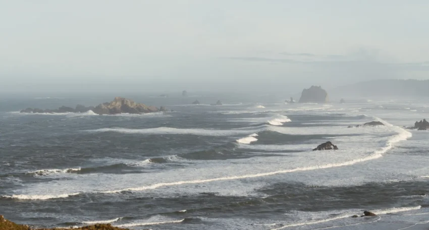 Vista panorámica del océano con olas rompiendo sobre rocas, en un día nublado. Se aprecian diversas formaciones rocosas y una costa lejana, evocando la belleza natural de la región costera.