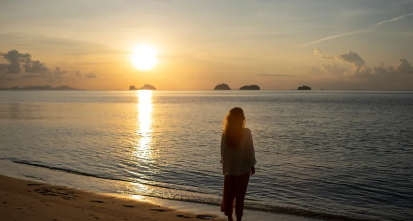 Mujer de pie en la playa durante el atardecer, contemplando el mar y las islas en el horizonte, con reflejos dorados en el agua.