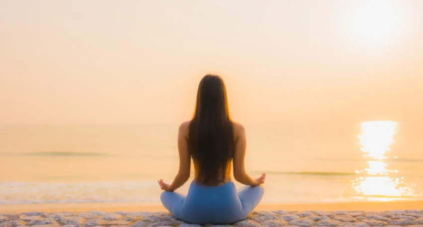 Mujer meditando en la playa al amanecer, sentada sobre una piedra, con vista al océano y el sol saliendo en el horizonte.